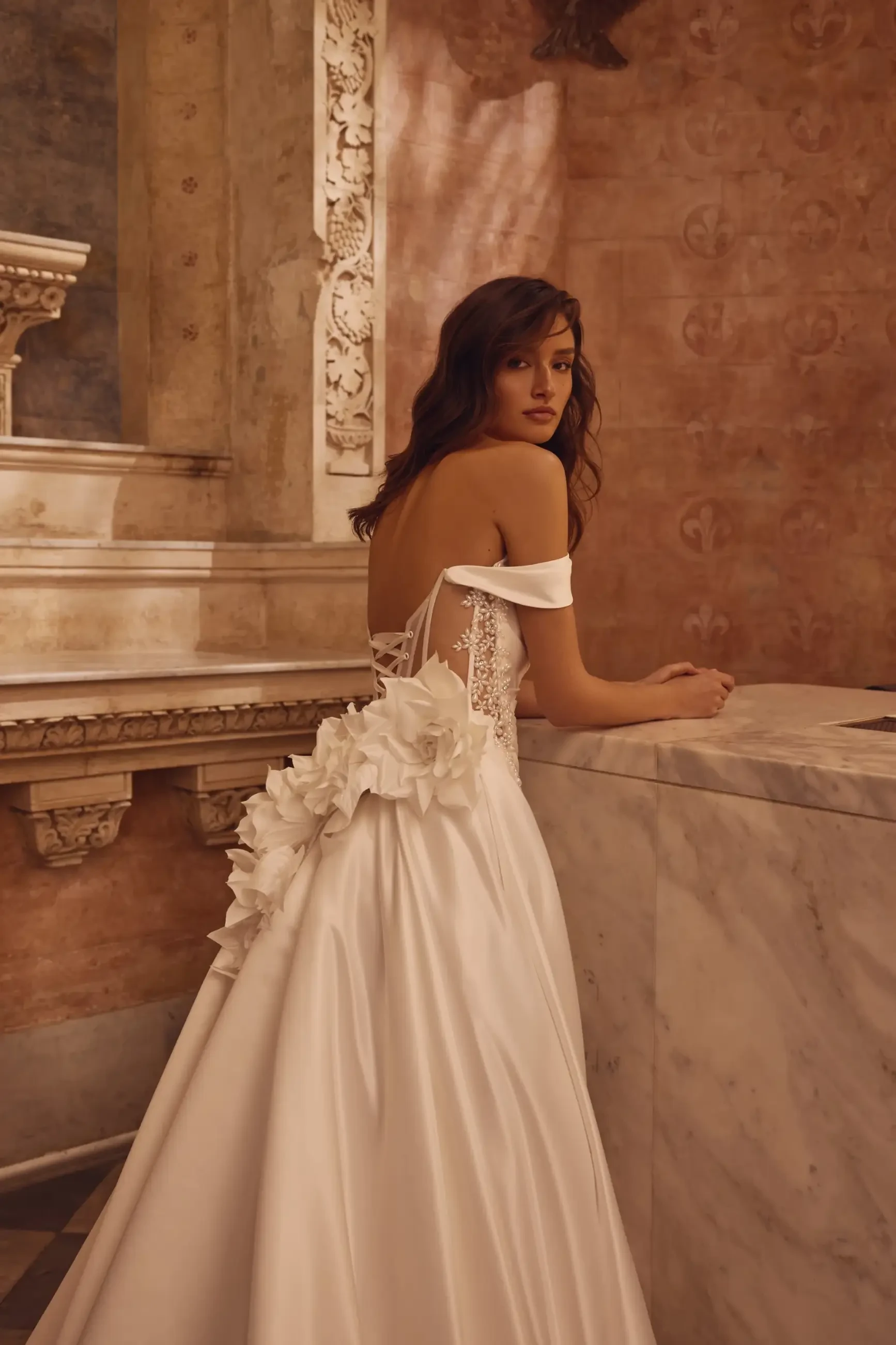 Model is wearing a satin A-line gown with off-the-shoulder draped straps, a pearl-embellished bodice, a thigh-high slit, and signature floral details.