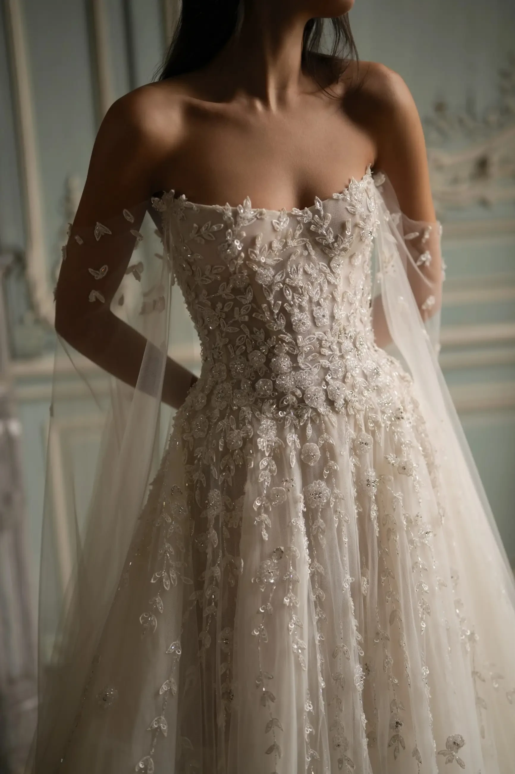 Model is wearing a voluminous ball gown with a structured corset bodice and shimmering beaded leaf appliqués, paired with flowing off-the-shoulder cape sleeves.