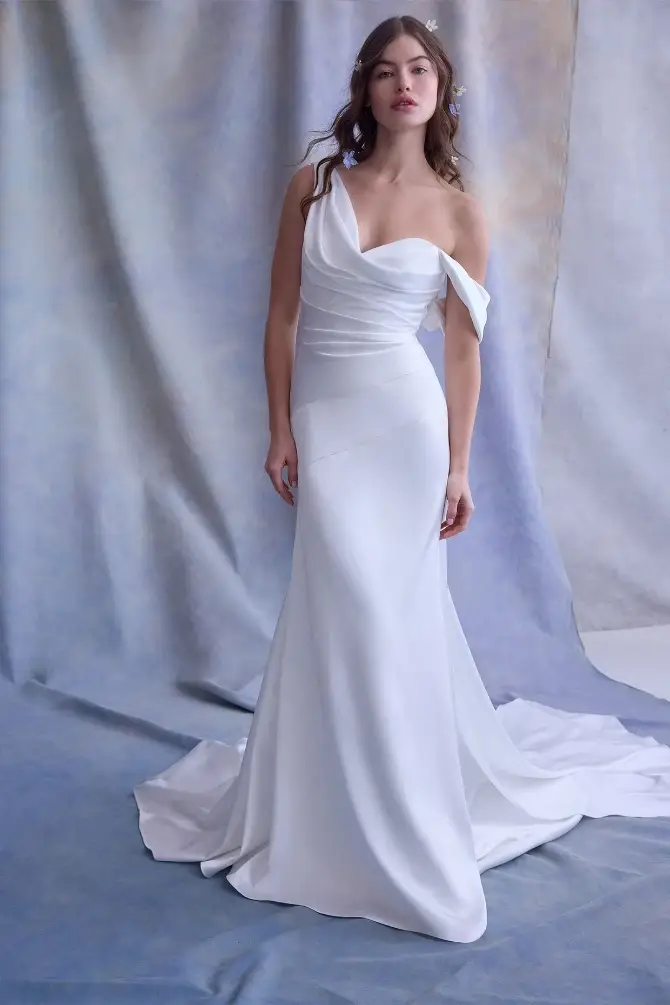 A woman in an elegant, off-the-shoulder white wedding gown with a flowing train stands against a soft, pastel-toned background, conveying grace and beauty.