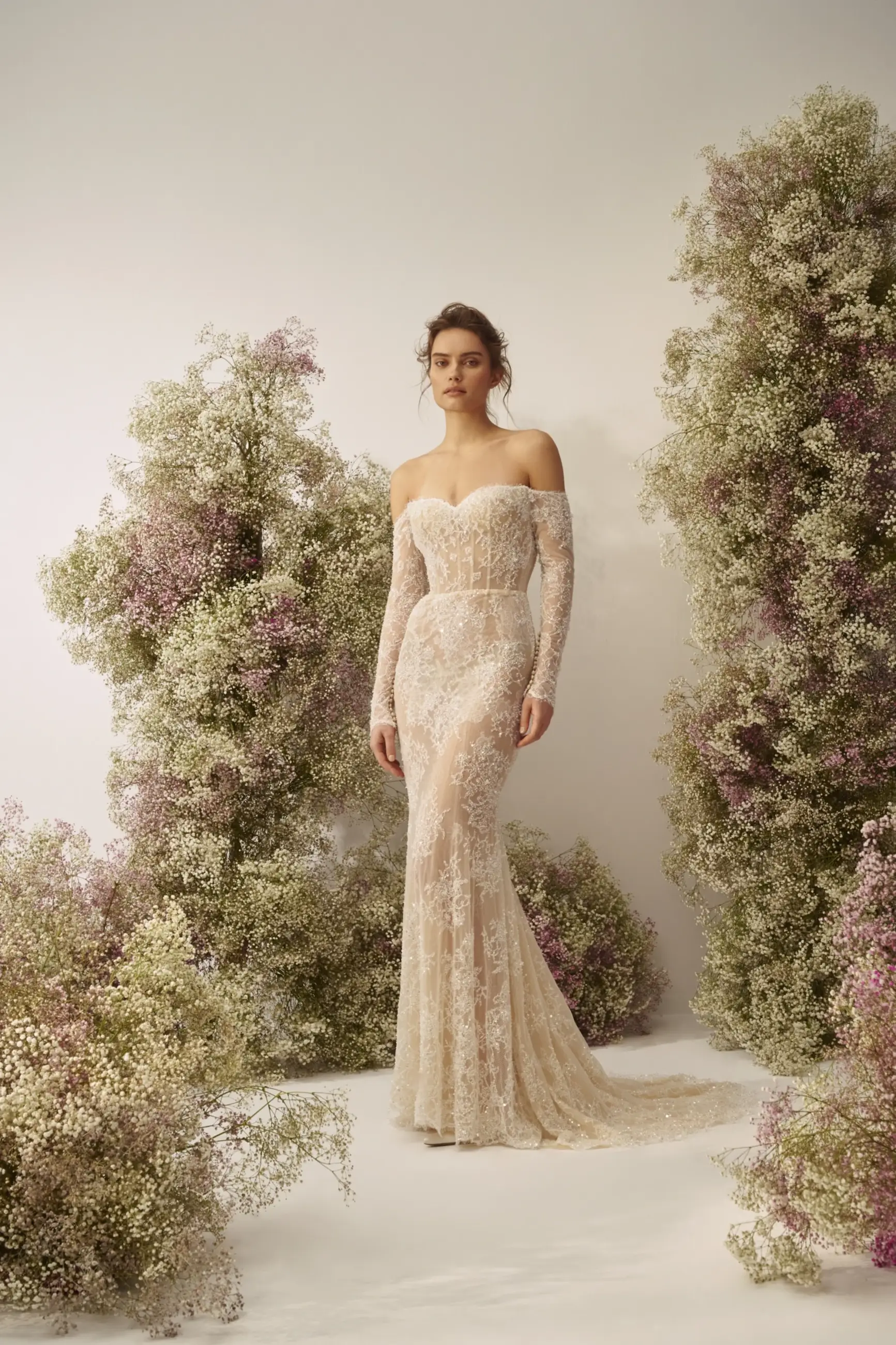 Model is wearing a fitted lace gown with an off-the-shoulder sweetheart neckline, beaded long sleeves, and a detachable silk overskirt.