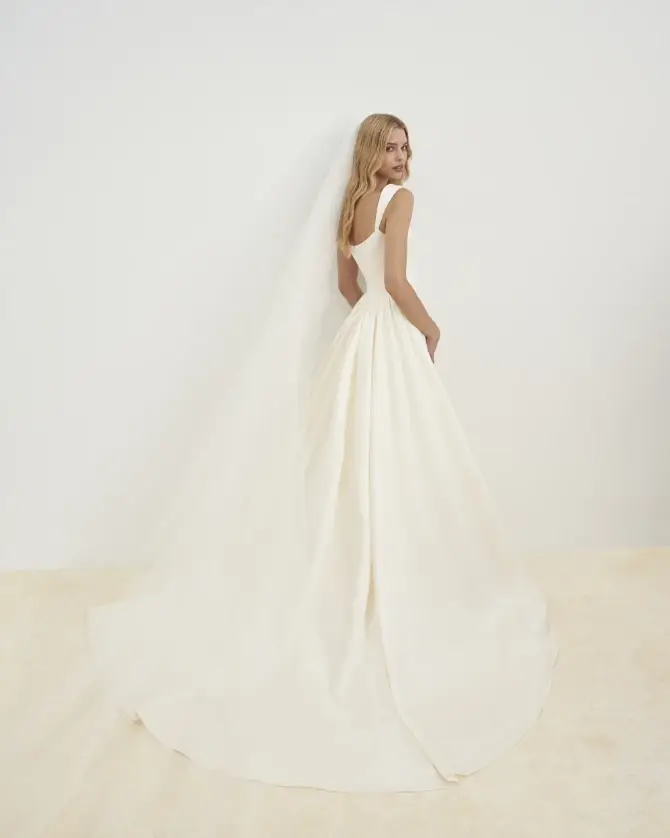 Model is wearing a fit-and-flare satin dress with tapered straps, a square neckline, and an exaggerated pleated panel train crafted from Silk Mikado