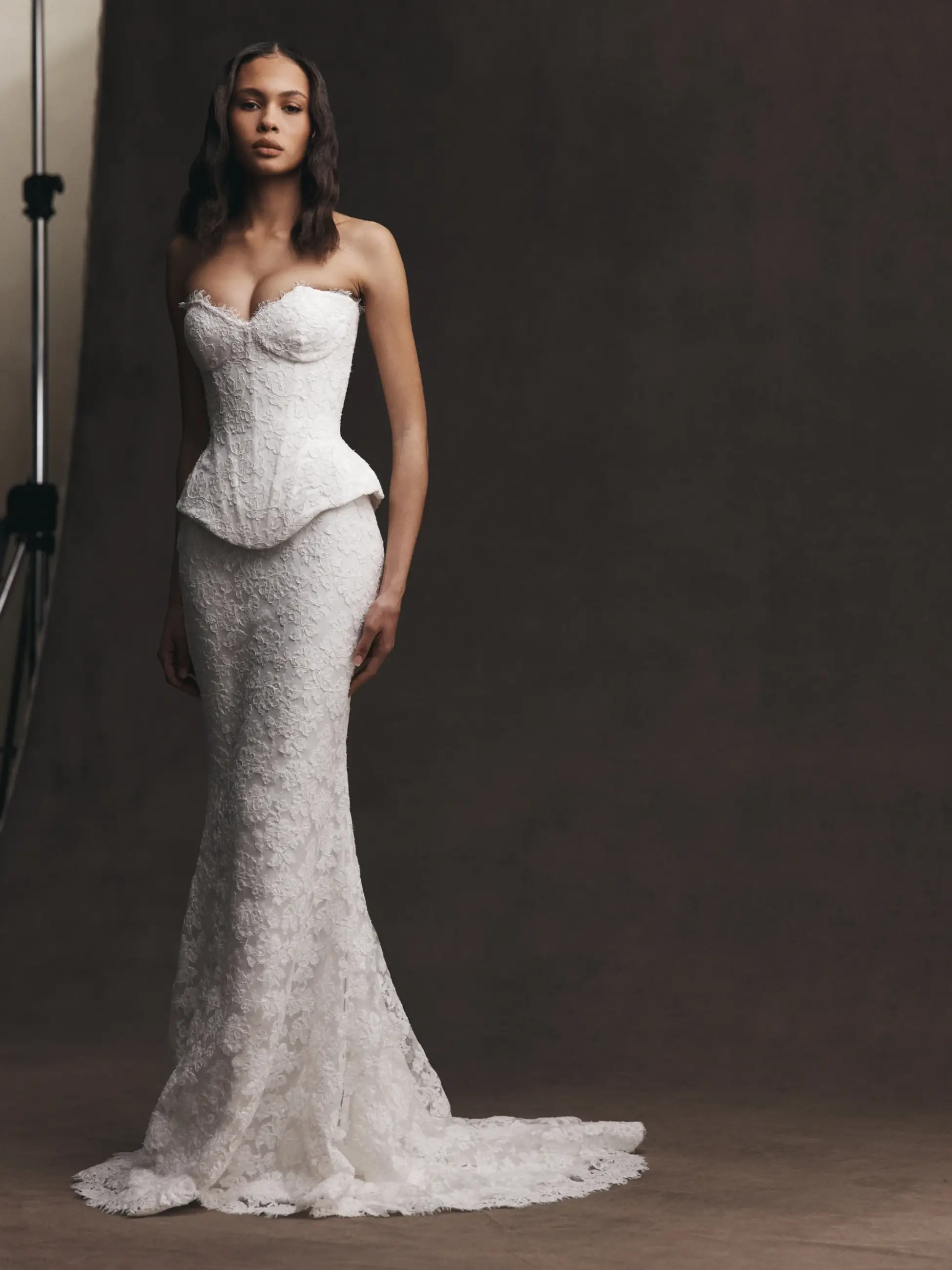 A model wearing a strapless white lace gown with a fitted bodice and flared bottom, standing against a neutral background.