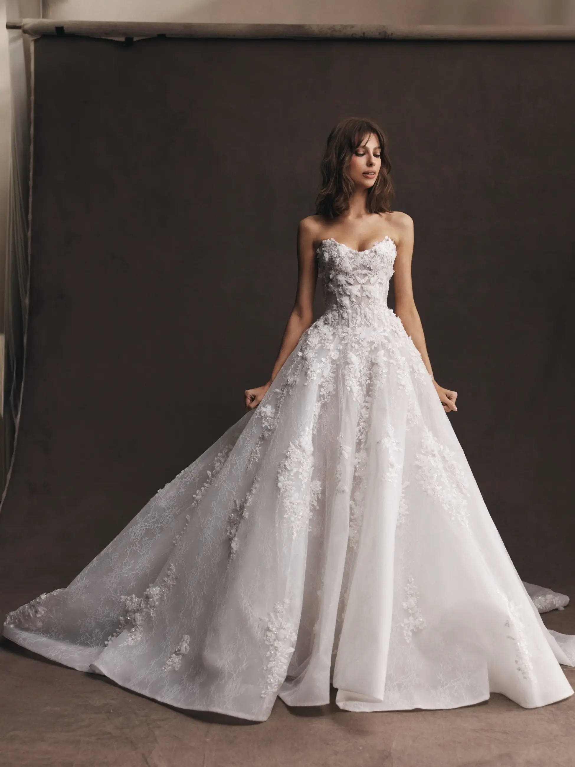 A woman in a strapless white wedding gown with intricate floral details, standing elegantly against a dark background.
