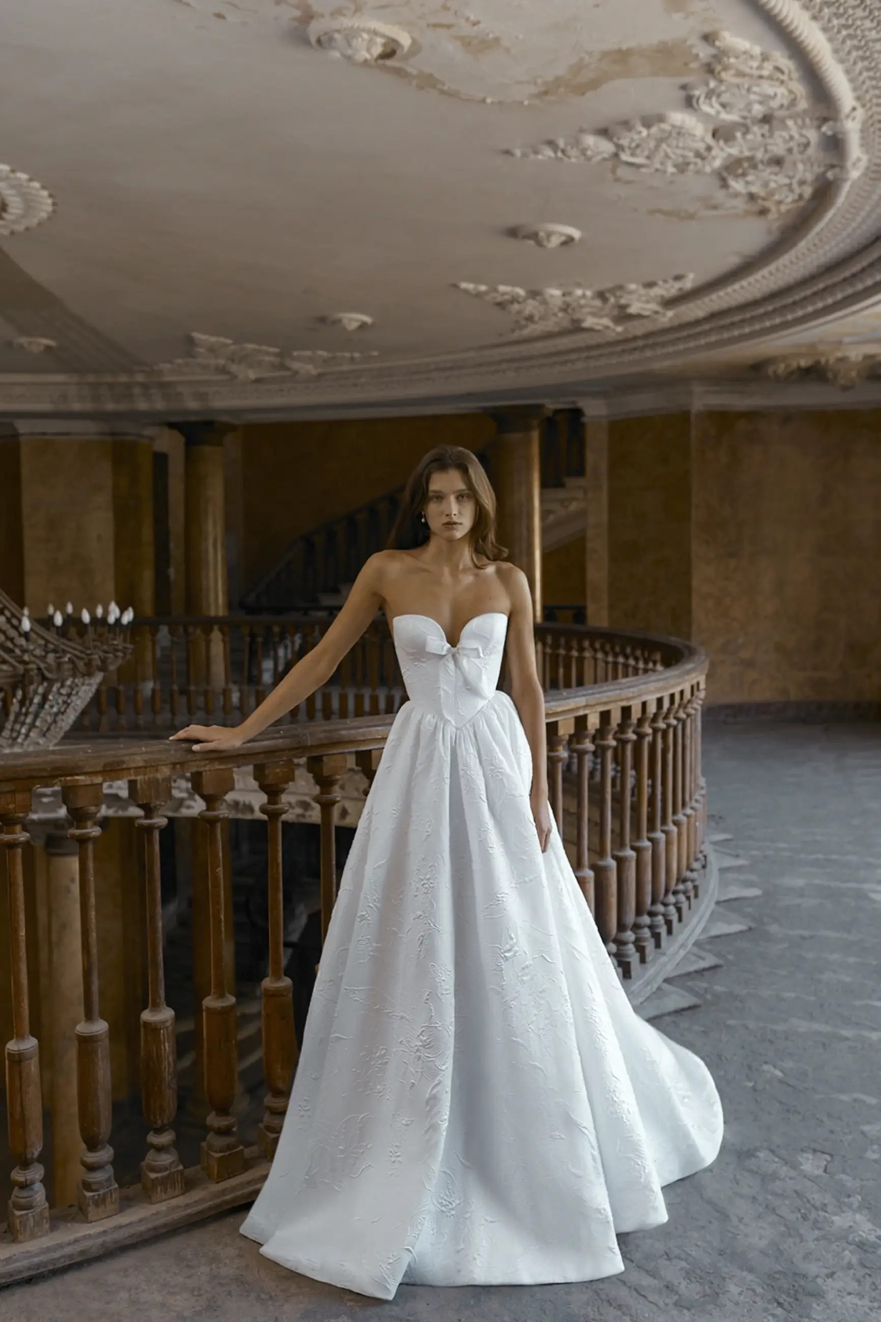  Elegant woman in a strapless white gown stands by a detailed wooden railing. The ornate ceiling above adds a vintage, romantic ambiance.