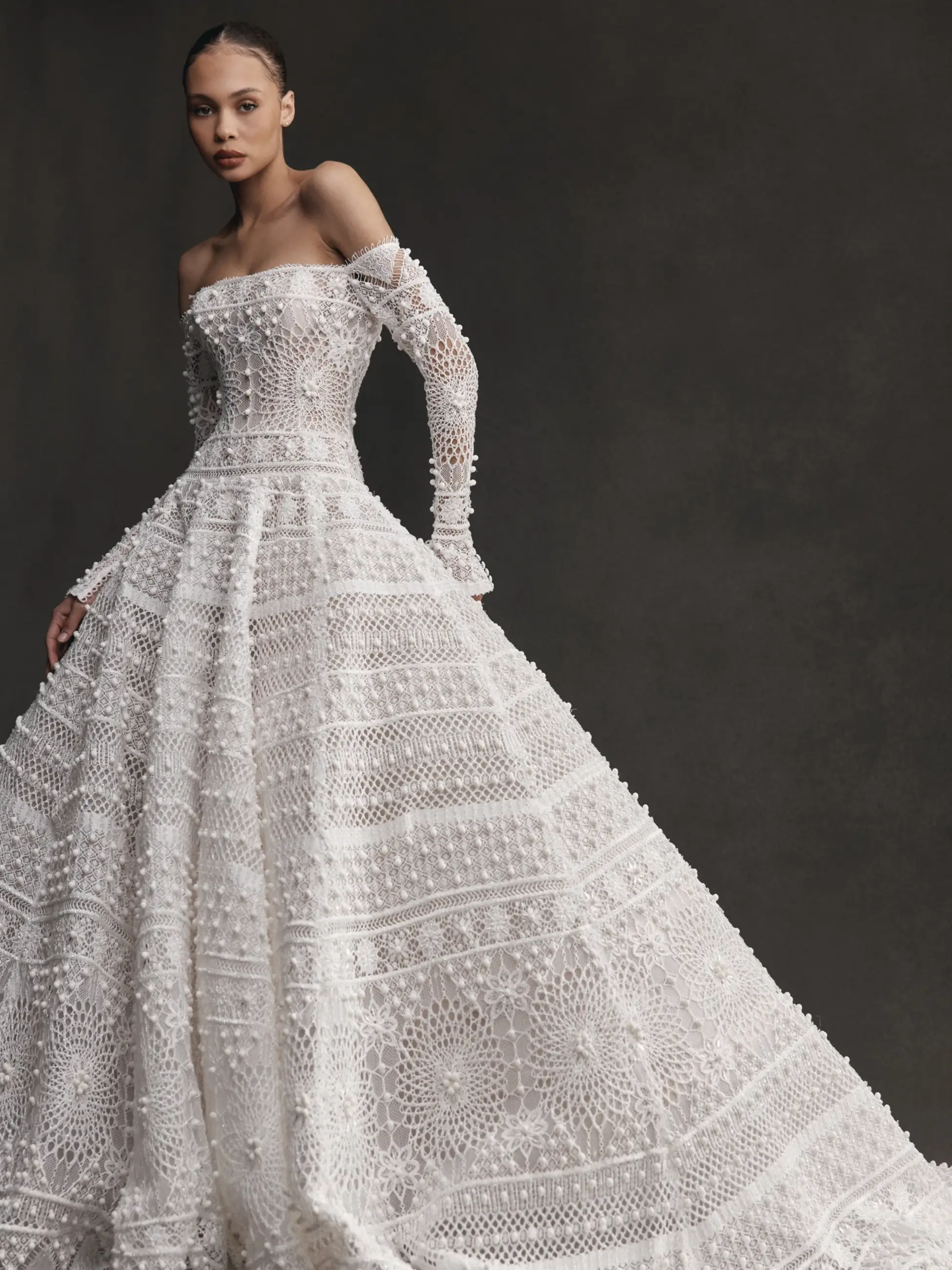 A model wearing an elegant white gown with intricate lace details and long, fitted sleeves. The dress has a full skirt and a structured bodice, creating a dramatic silhouette against a neutral backdrop.