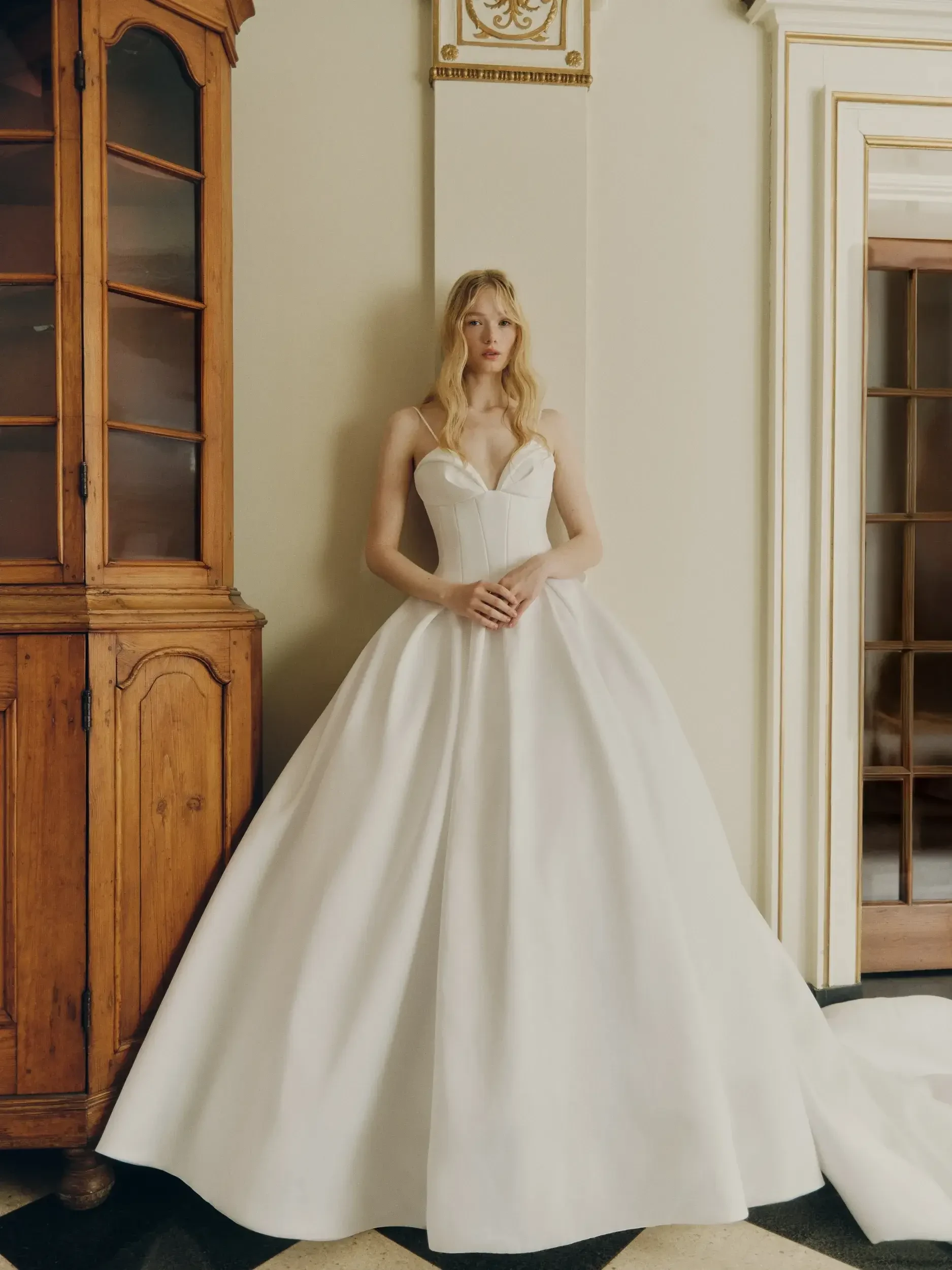 A woman in an elegant, strapless white ball gown stands in a room with vintage decor. She leans against a wooden cabinet, exuding a calm and poised demeanor.