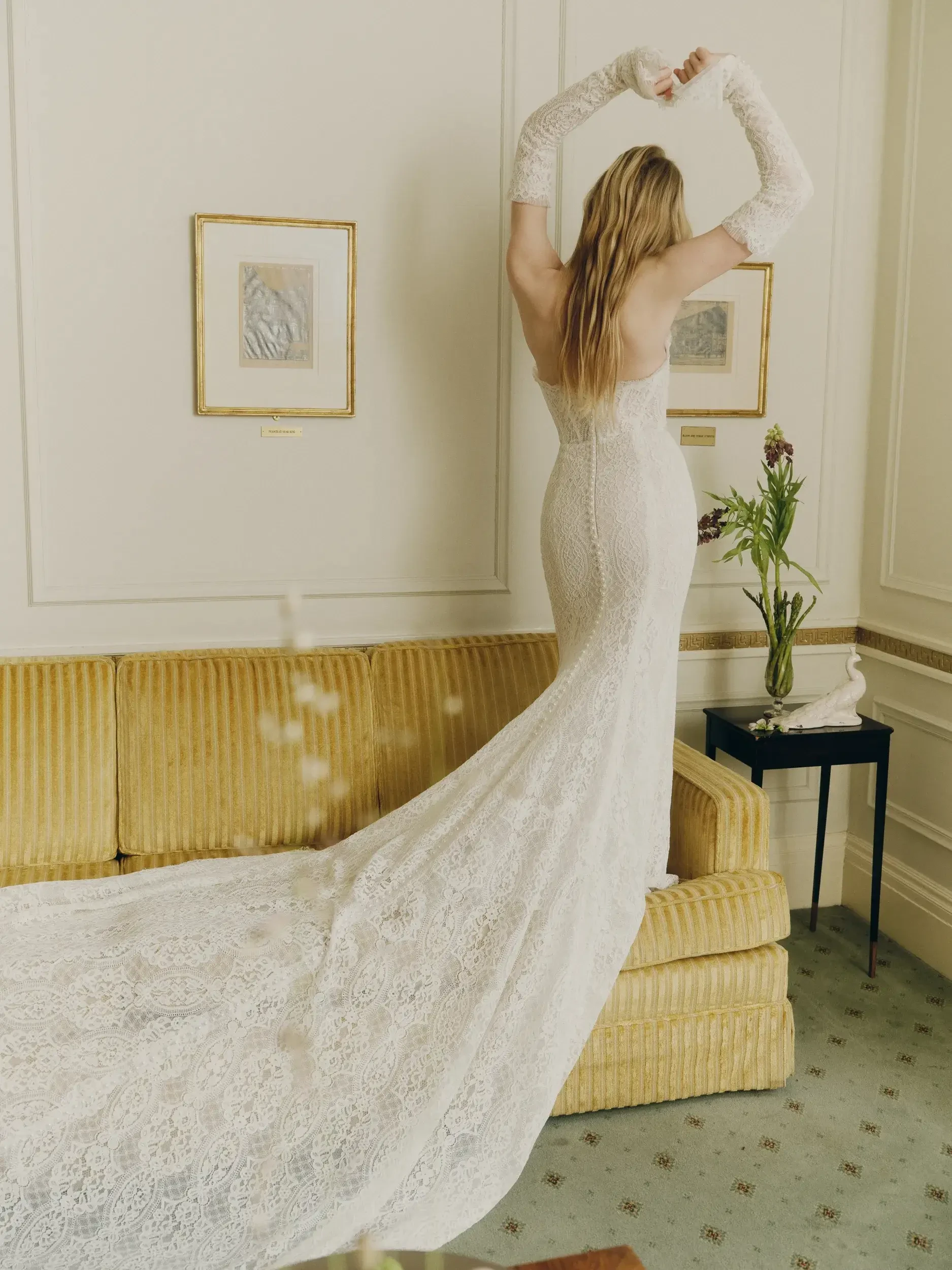 A woman in an elegant, lace wedding dress stands on a mustard-colored sofa, facing away. Her arms are raised, and the dress has a long train. The room is decorated with framed art, a small side table with flowers, and a sculpture, conveying a classic, sophisticated atmosphere.