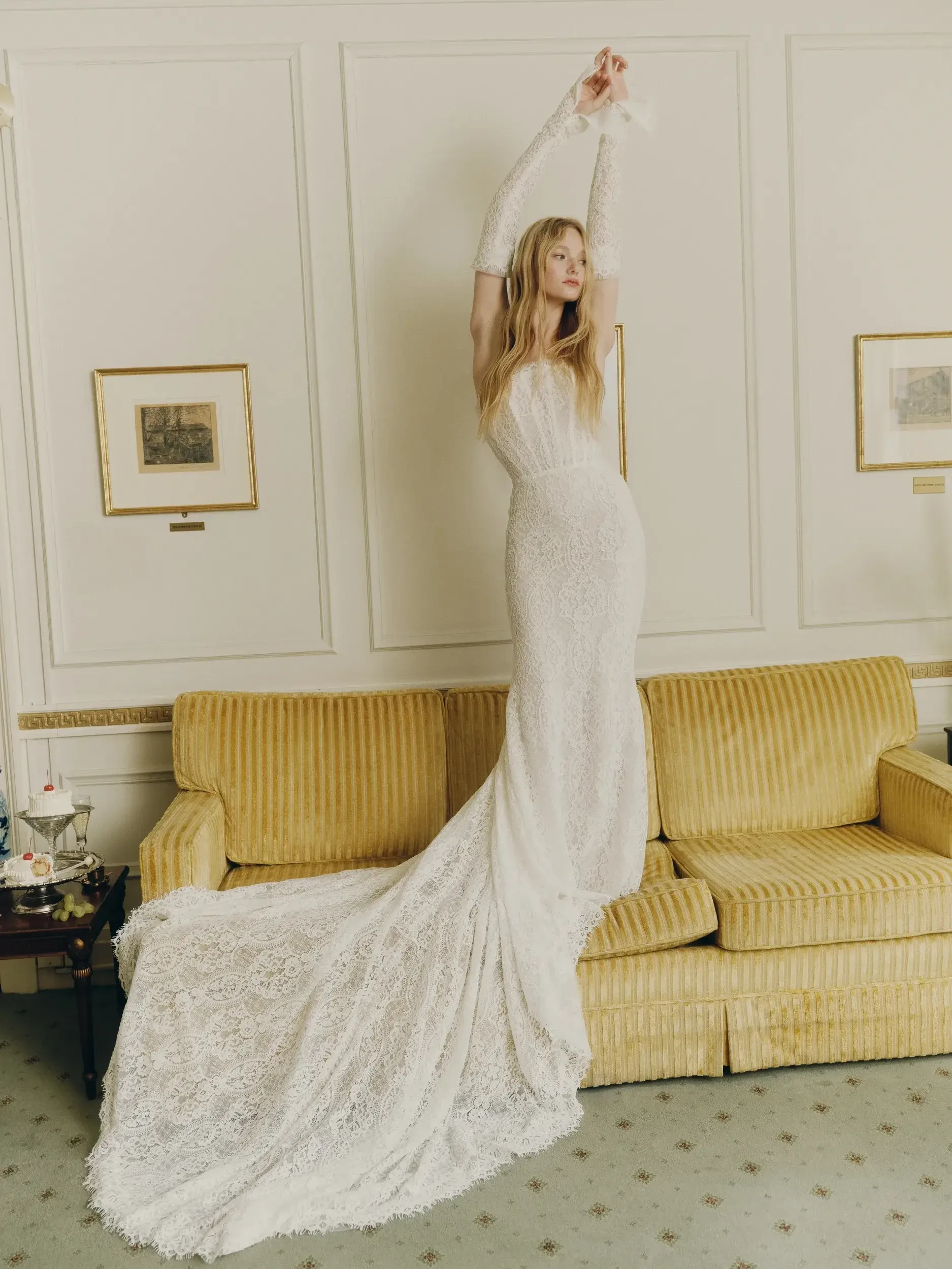 A woman in a long, elegant lace wedding gown poses with arms raised on a yellow sofa in a classic room, conveying grace and sophistication.