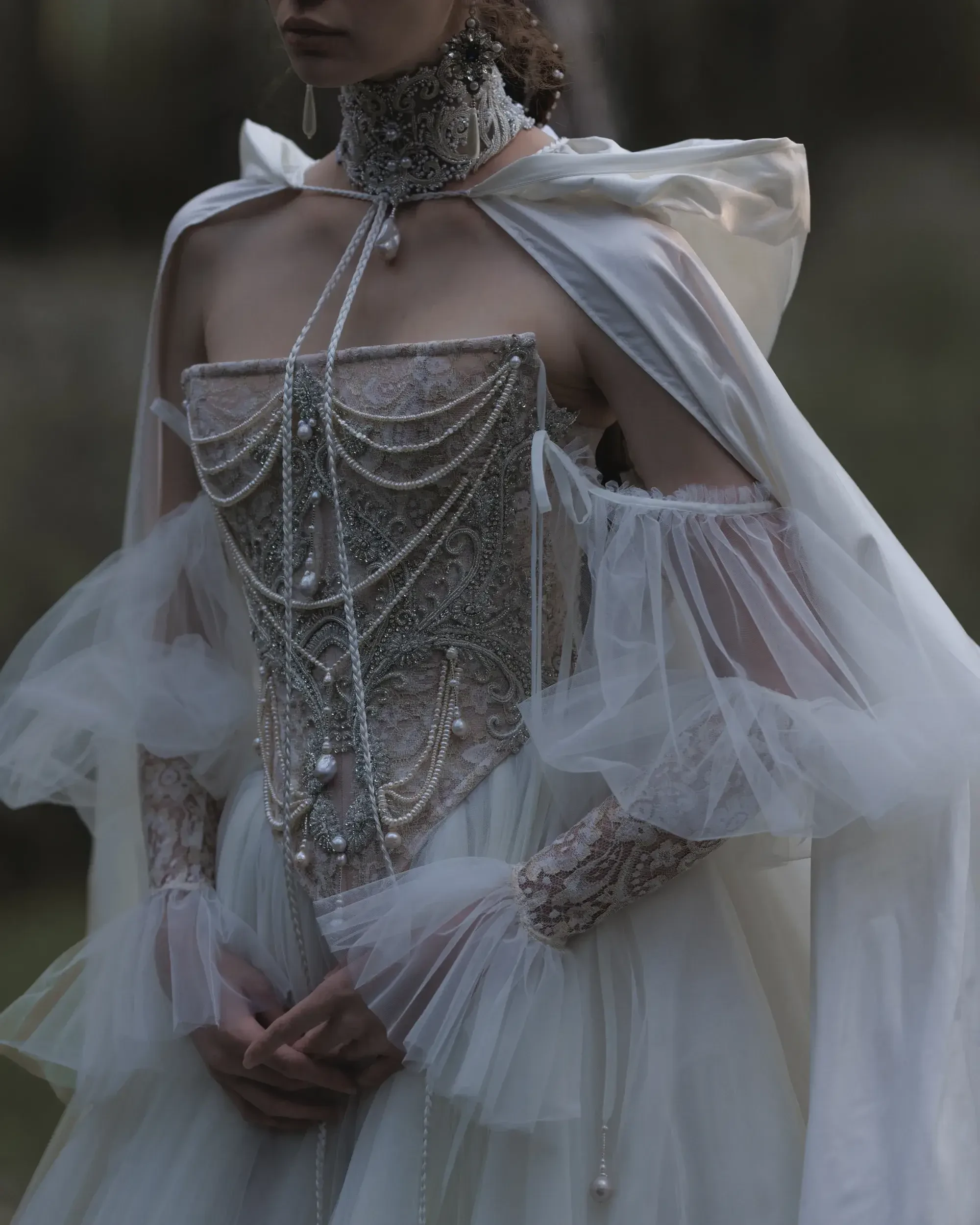 A woman wears an elegant, intricately beaded corset with sheer, flowing sleeves and a matching cape. The ambiance is dreamy and ethereal.