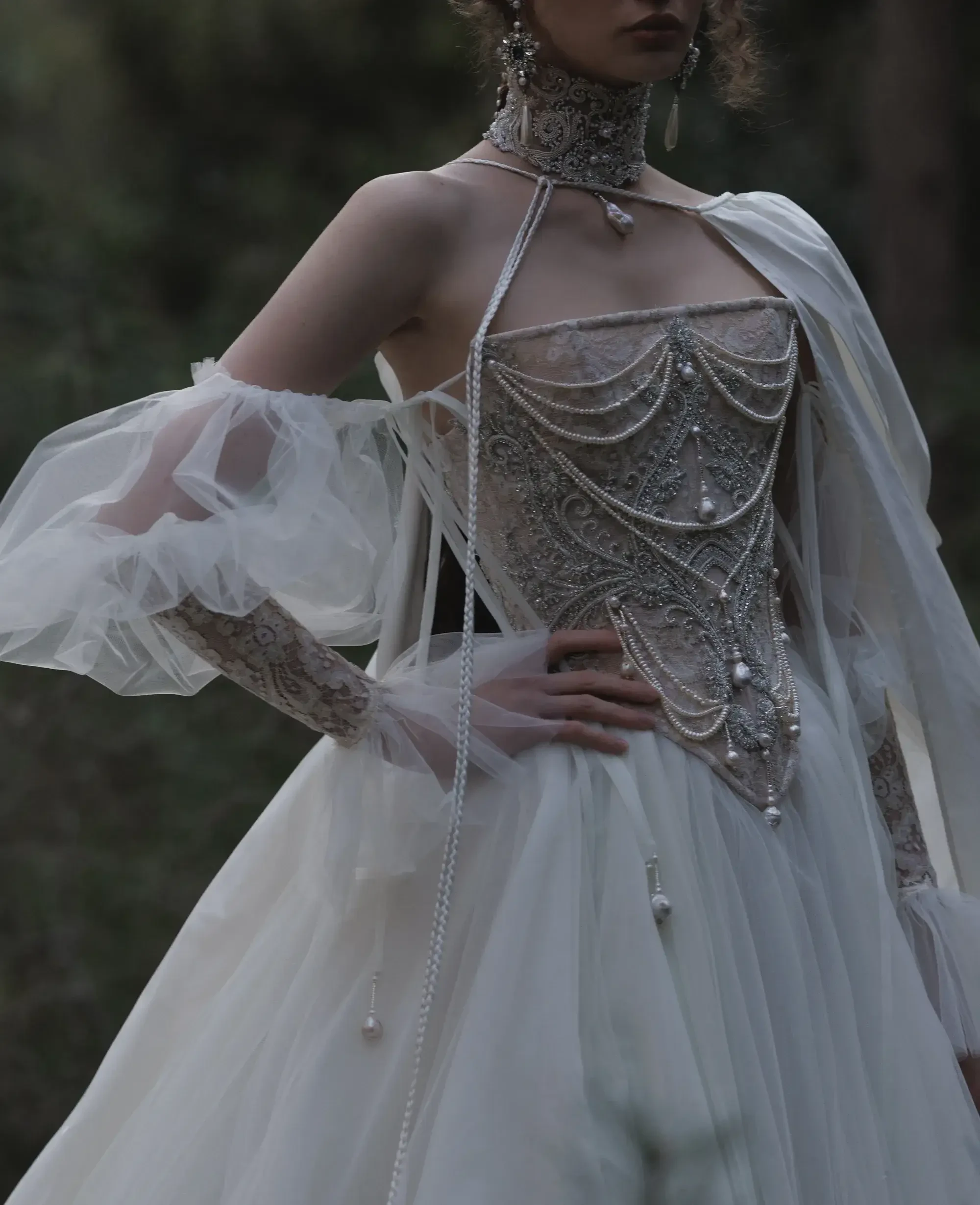 A model wearing an intricately designed wedding gown with puffed sleeves and ornate embellishments, standing amidst a natural setting.