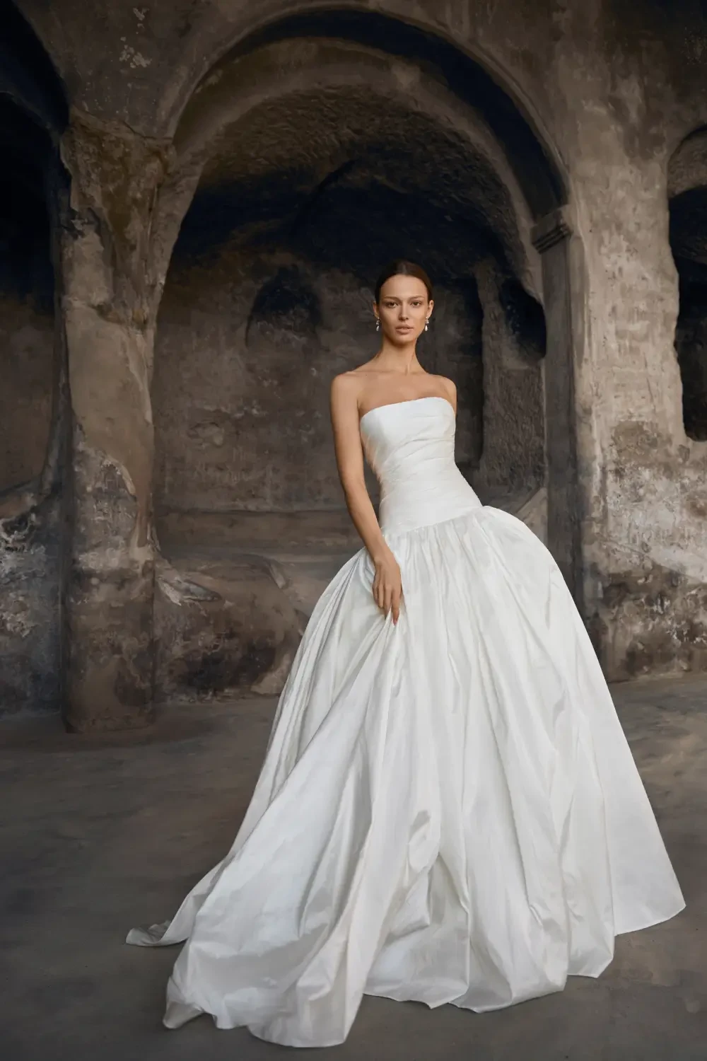 A model wearing a strapless white wedding gown stands in an ancient-looking architectural setting with stone arches.