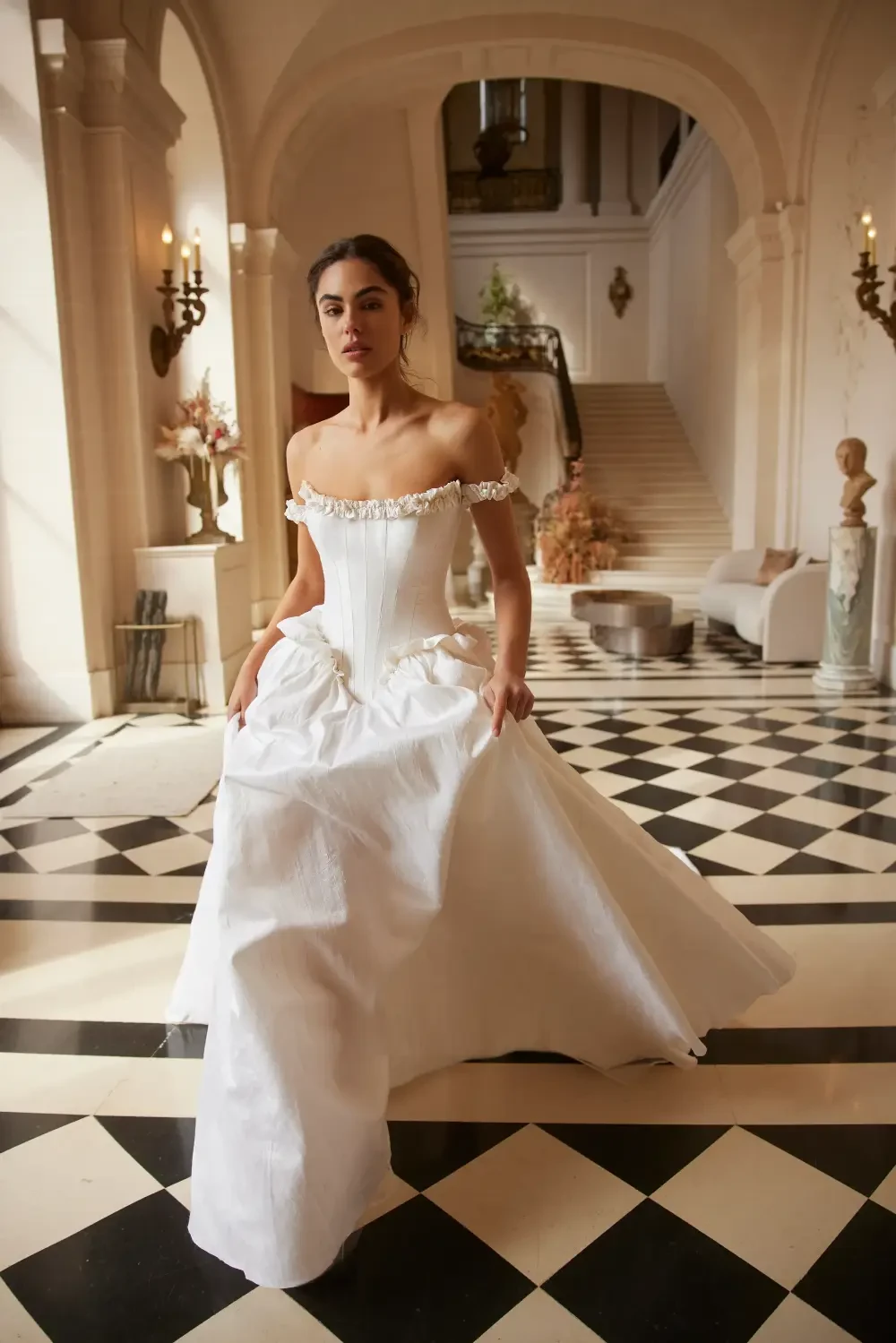 A woman in a white off-the-shoulder gown poses in a grand hall with a black and white checkered floor and elegant decor.