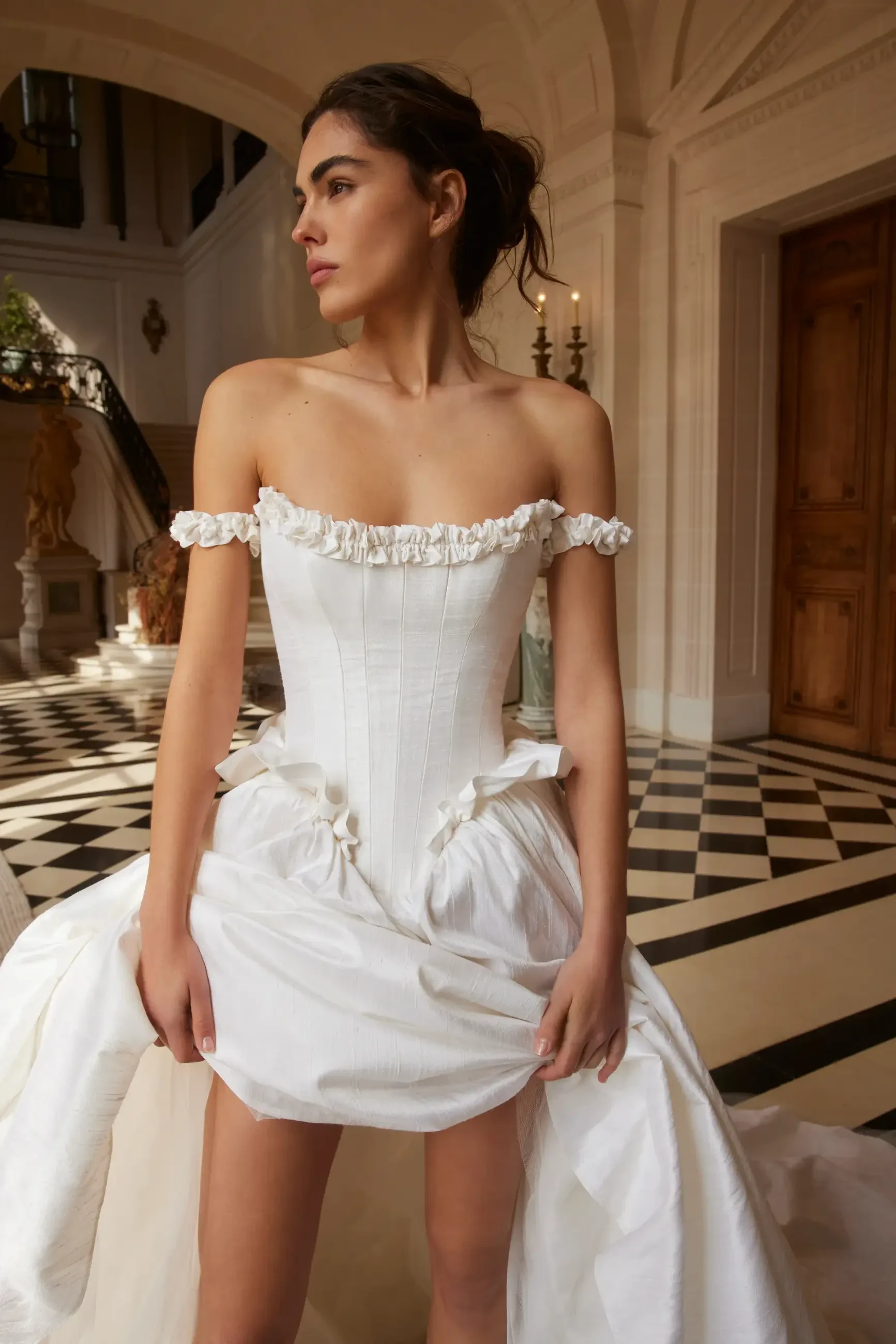 A woman in a strapless white gown stands in an elegant, sunlit hallway with checkerboard flooring, looking to the side. The scene exudes sophistication.