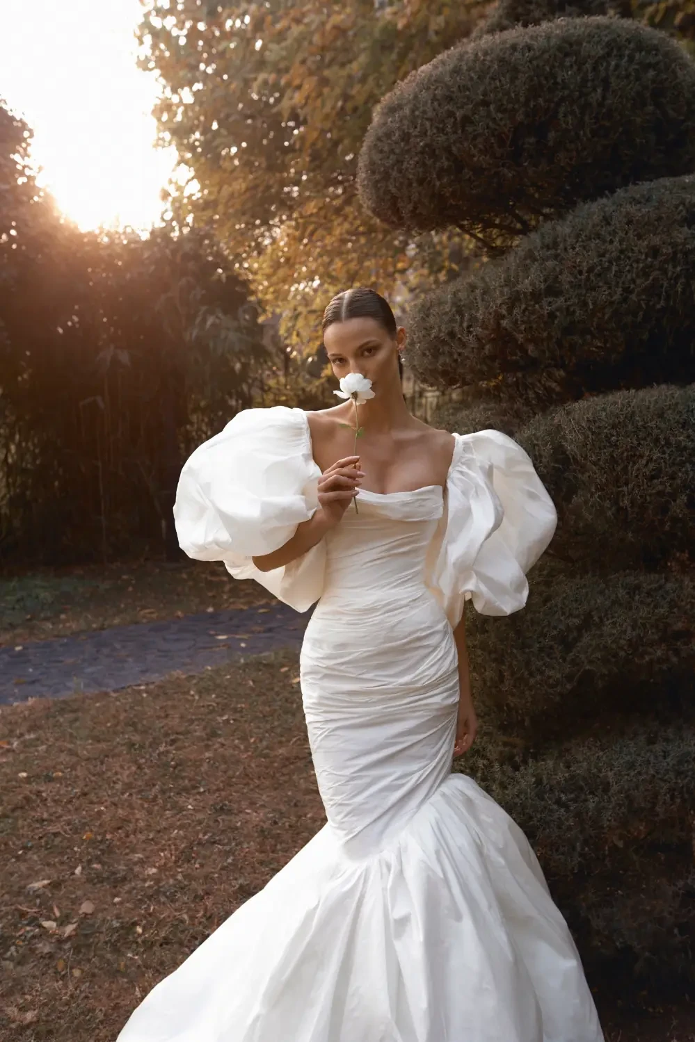 A model wearing a white gown with large puff sleeves, holding a flower, standing in a garden setting with trees in the background.