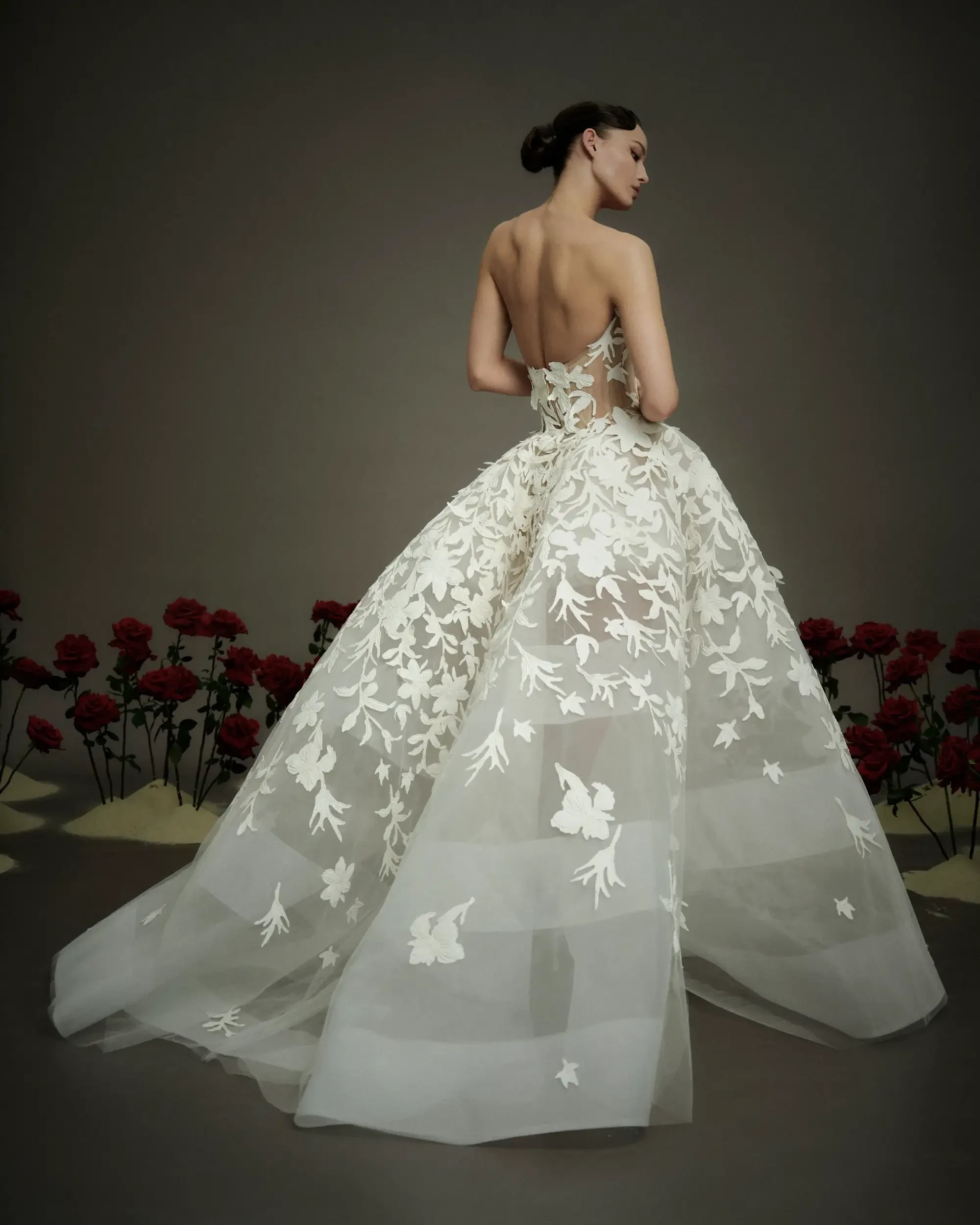 A woman in a strapless, backless gown adorned with floral lace stands gracefully. Red roses surround her, creating a romantic, elegant ambiance.