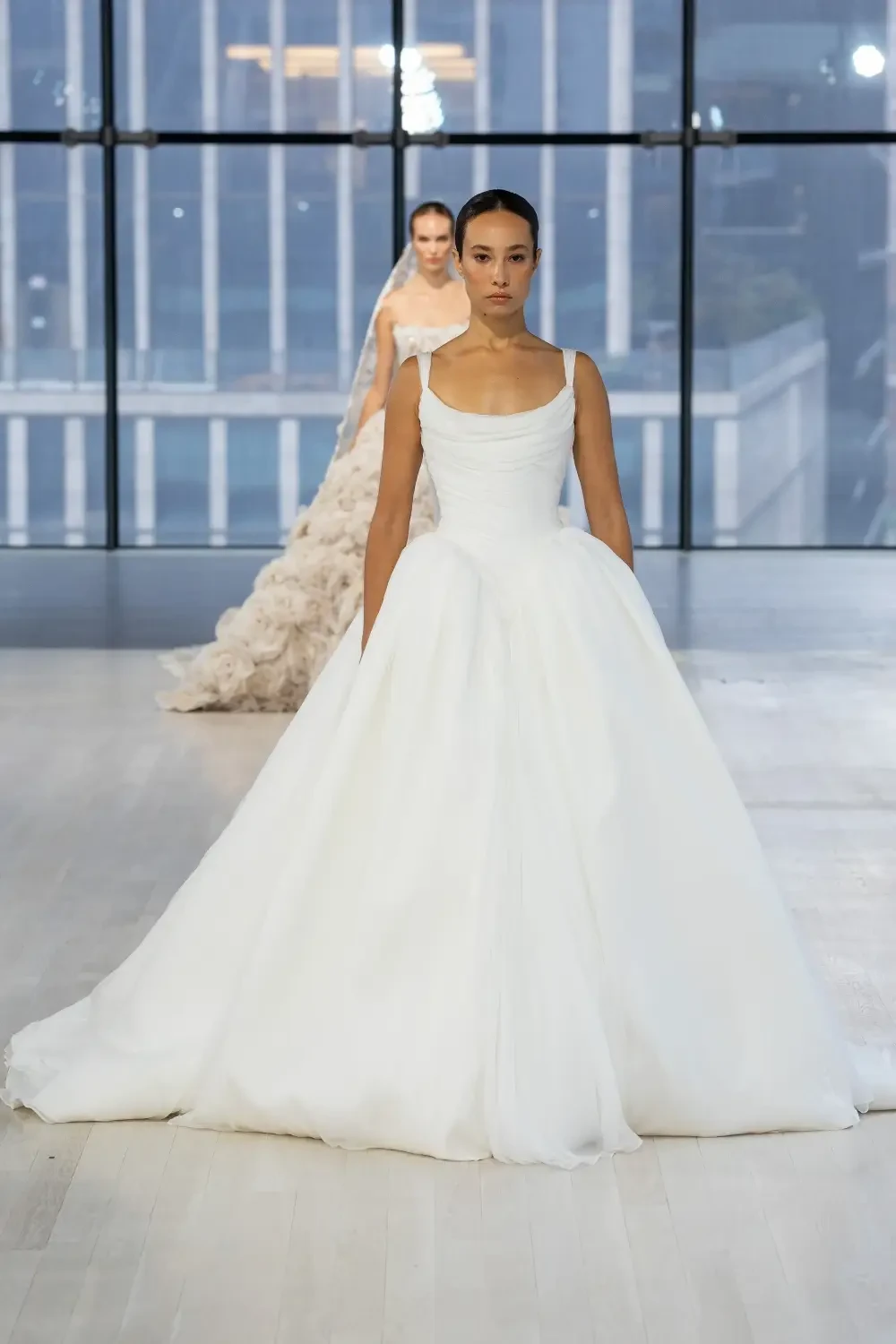 A model wearing a white gown walks down a runway, with another model in a gown in the background. The setting is an indoor venue with large windows.