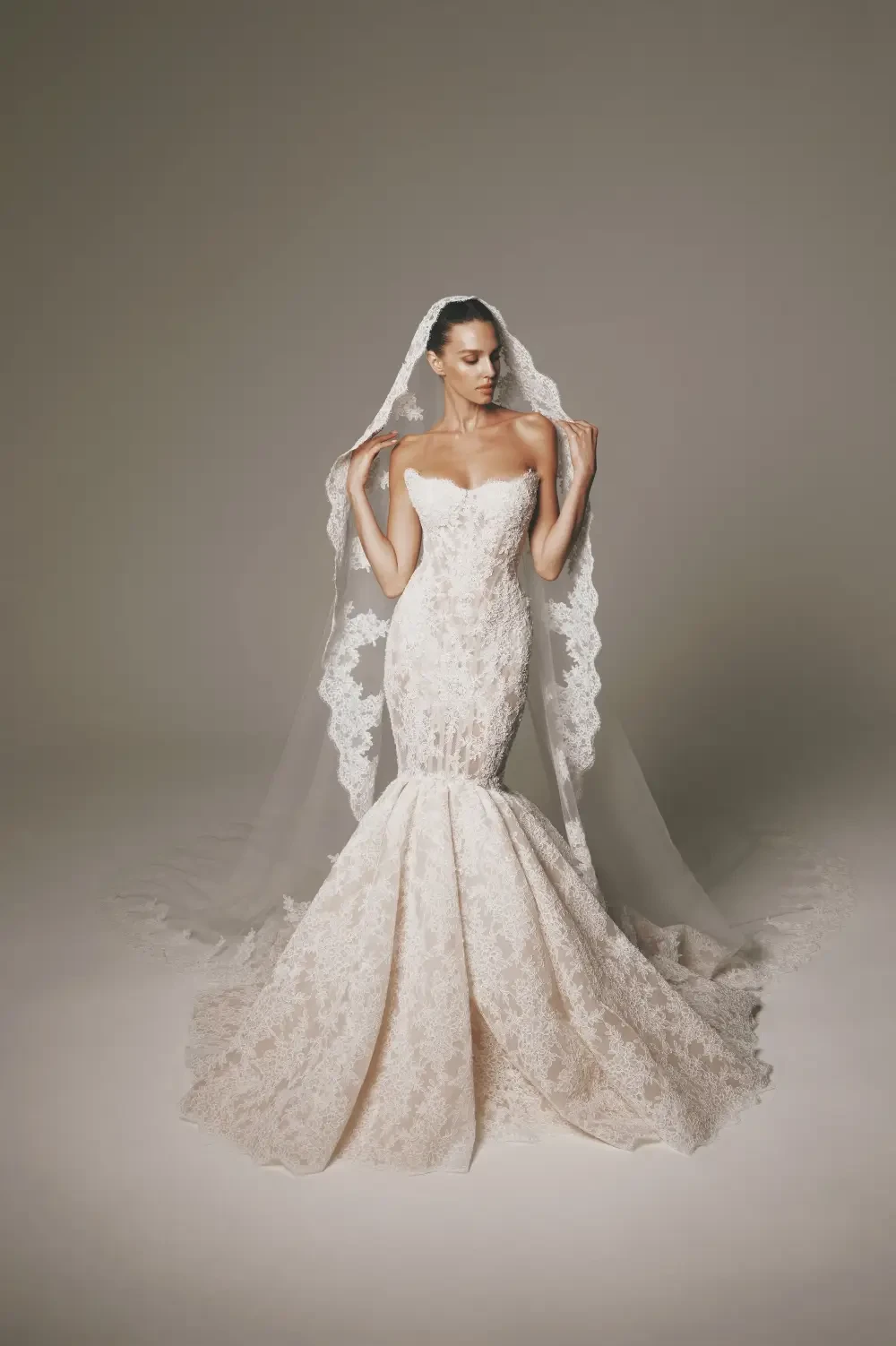 A bride in a strapless, mermaid-style wedding gown with intricate lace detail, holding a long veil.