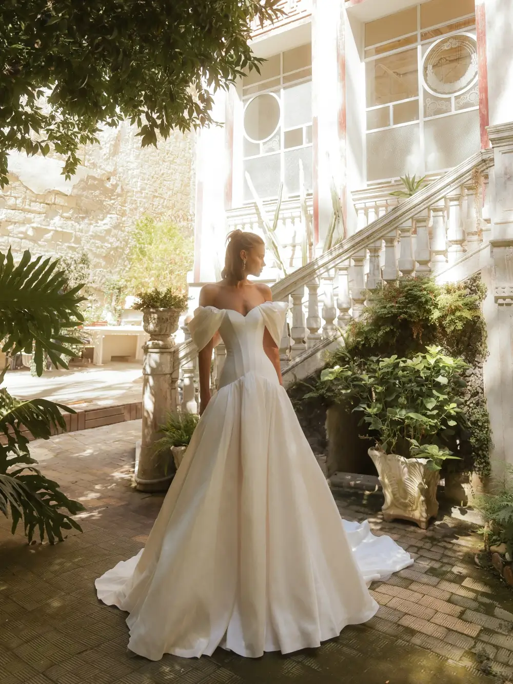 A woman in a white gown stands elegantly in a garden surrounded by greenery and classical architecture.