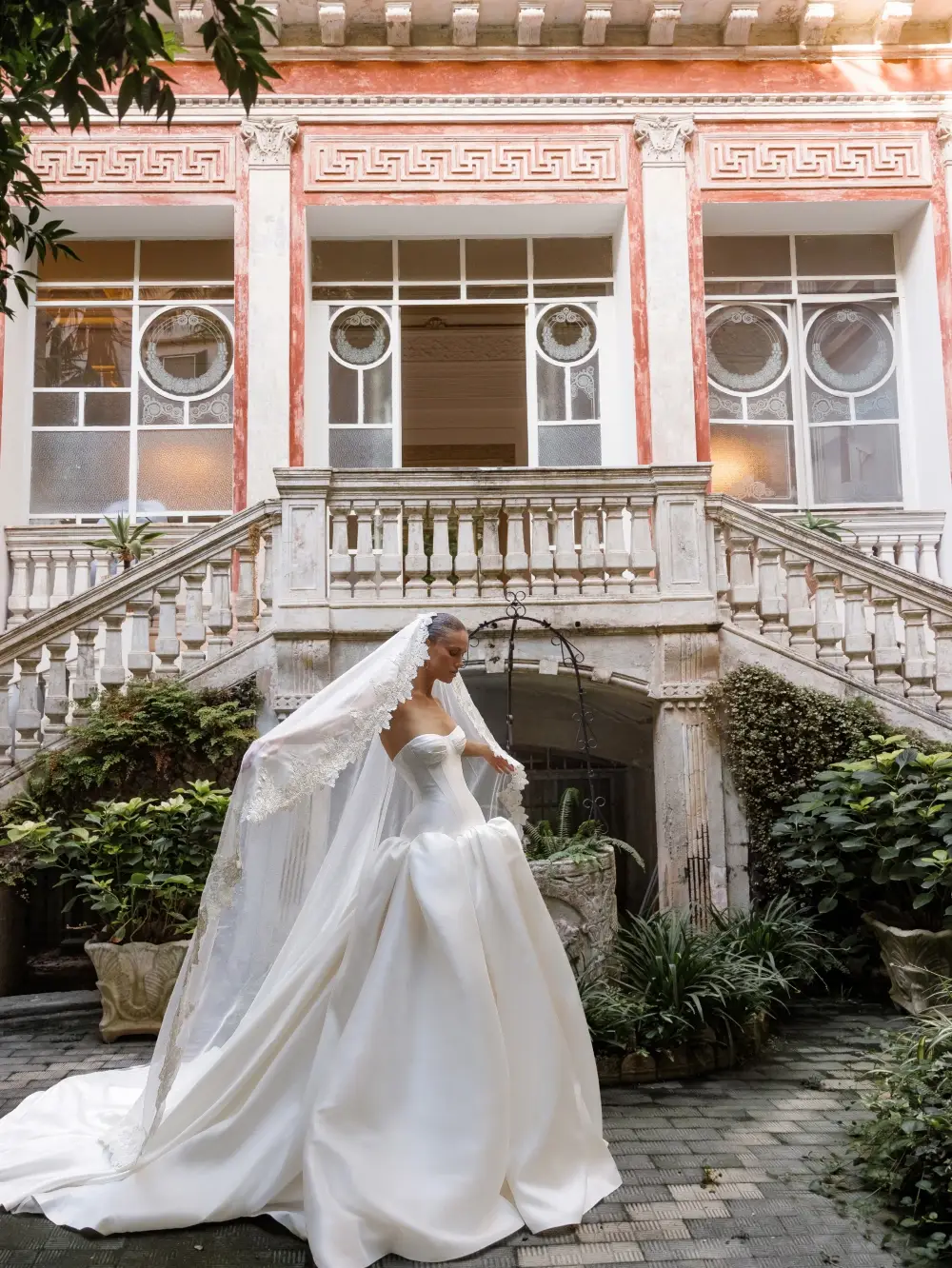 A bride in a white gown and veil stands in a beautifully landscaped courtyard with a historic building in the background.