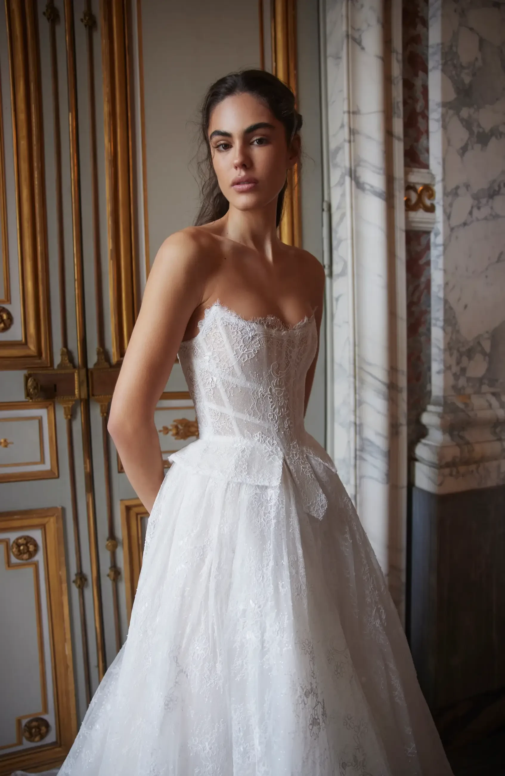 A woman in a strapless white wedding dress stands in front of ornate gold and marble decor.